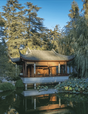 A traditional Chinese pavilion with a curved roof stands over a pond with lily pads, reflecting in the water and surrounded by trees.