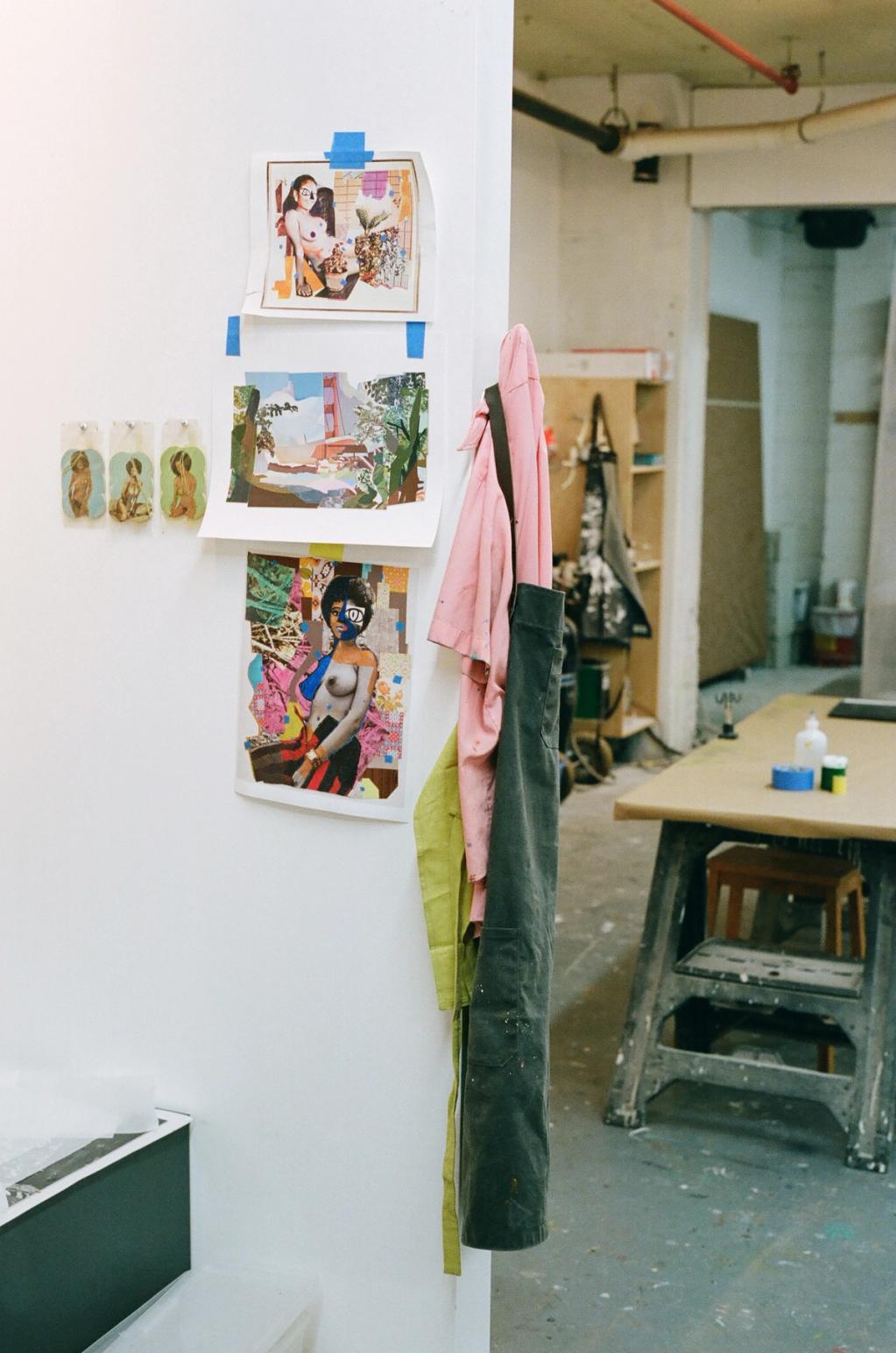 A white wall in an art studio displays three prints of figures and a landscape, taped to the wall. Aprons hang on the wall's edge, with a large work table and supplies visible in the background.