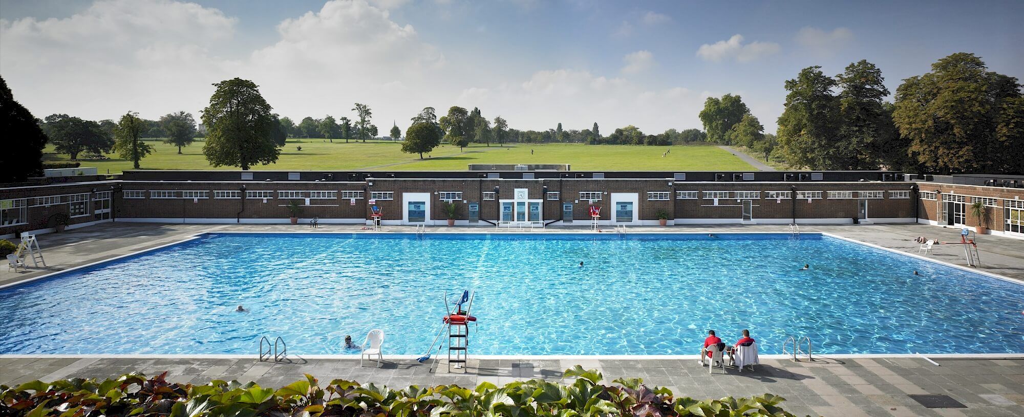 A large outdoor swimming pool with bathers, a facility building, and a green park in the background.
