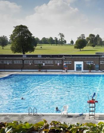 A large outdoor swimming pool with bathers, a facility building, and a green park in the background.