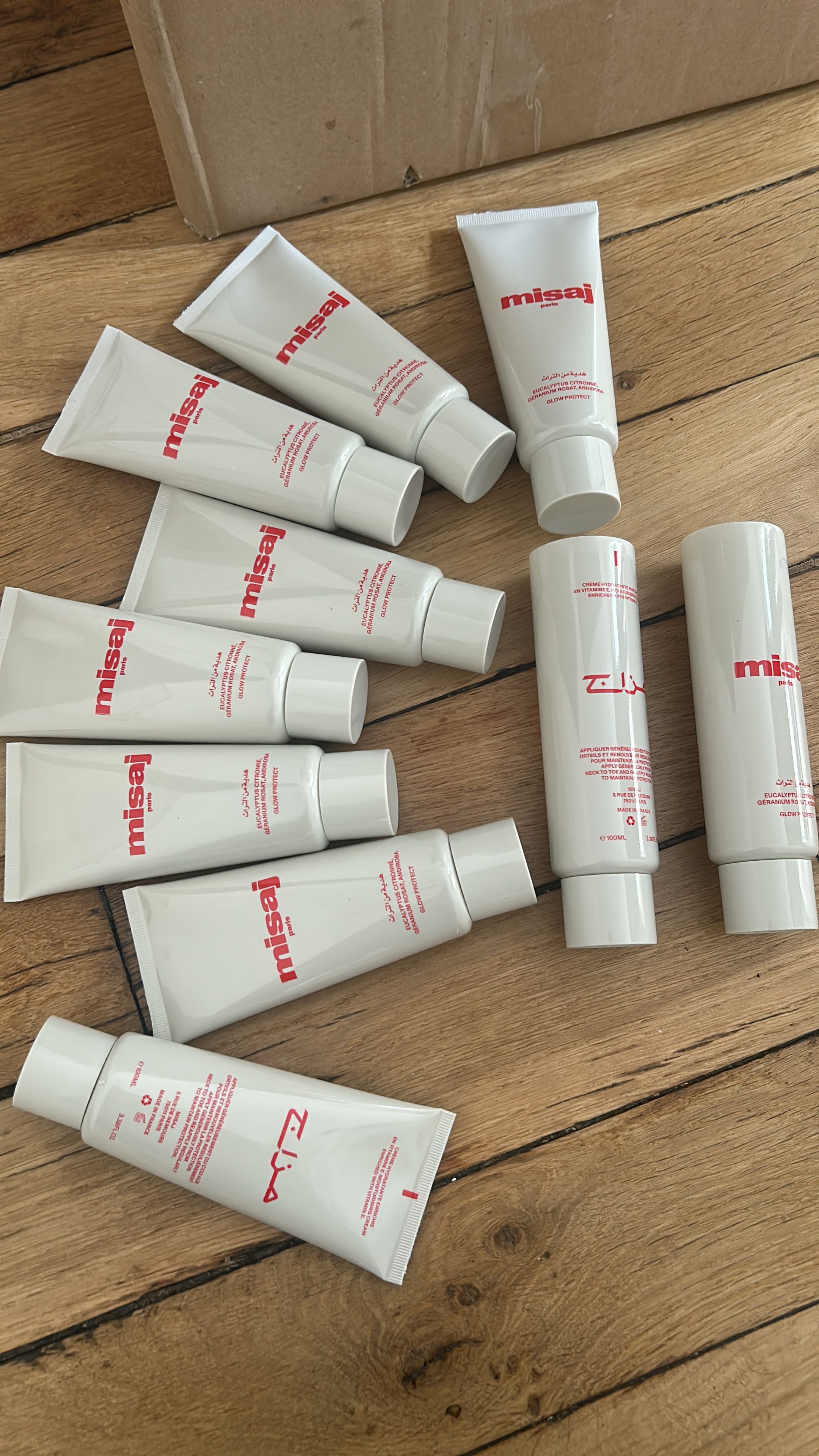 Multiple white tubes of Misaj Paris cream with red text, scattered on a wooden floor next to a cardboard box.