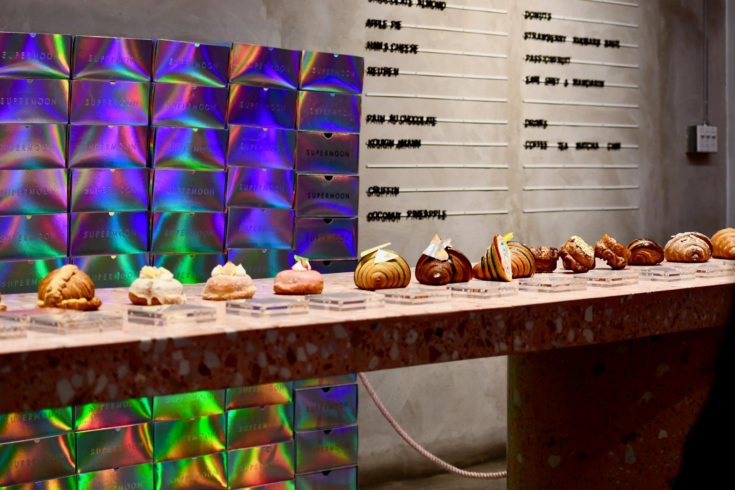 A patisserie display counter features various croissants and donuts, backed by a wall of iridescent "SUPERMOON" boxes and a menu board.