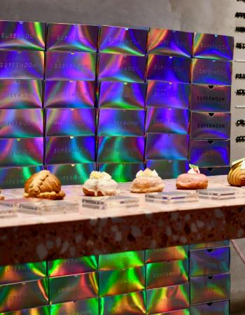 A patisserie display counter features various croissants and donuts, backed by a wall of iridescent "SUPERMOON" boxes and a menu board.