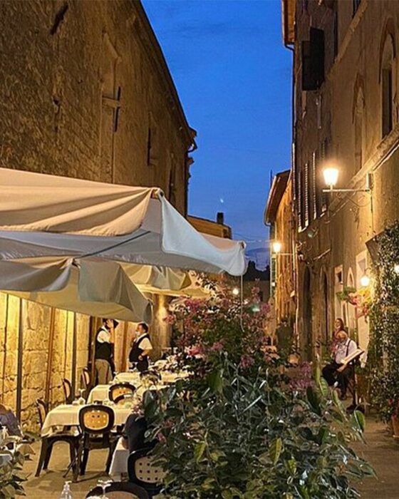 "An outdoor restaurant with tables under white umbrellas in a narrow, illuminated alley at dusk, with servers and a musician."