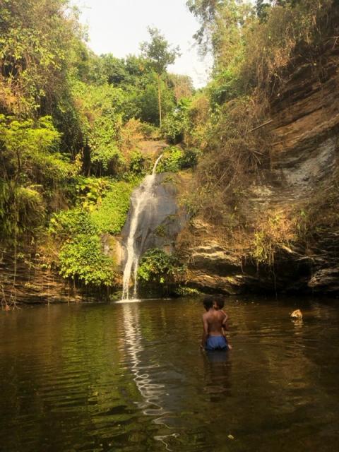Two people wade in a natural pool fed by a waterfall surrounded by lush green foliage and rocky cliffs.