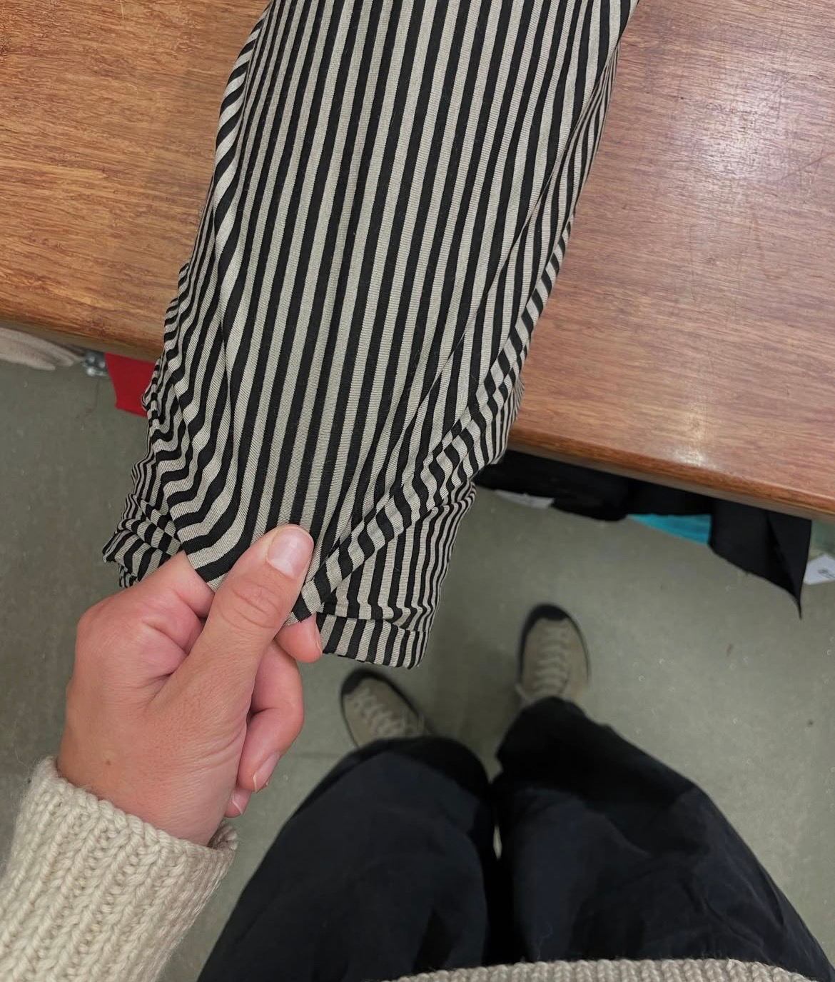 A hand holds up black and beige striped fabric, with legs in black pants and sneakers visible below.