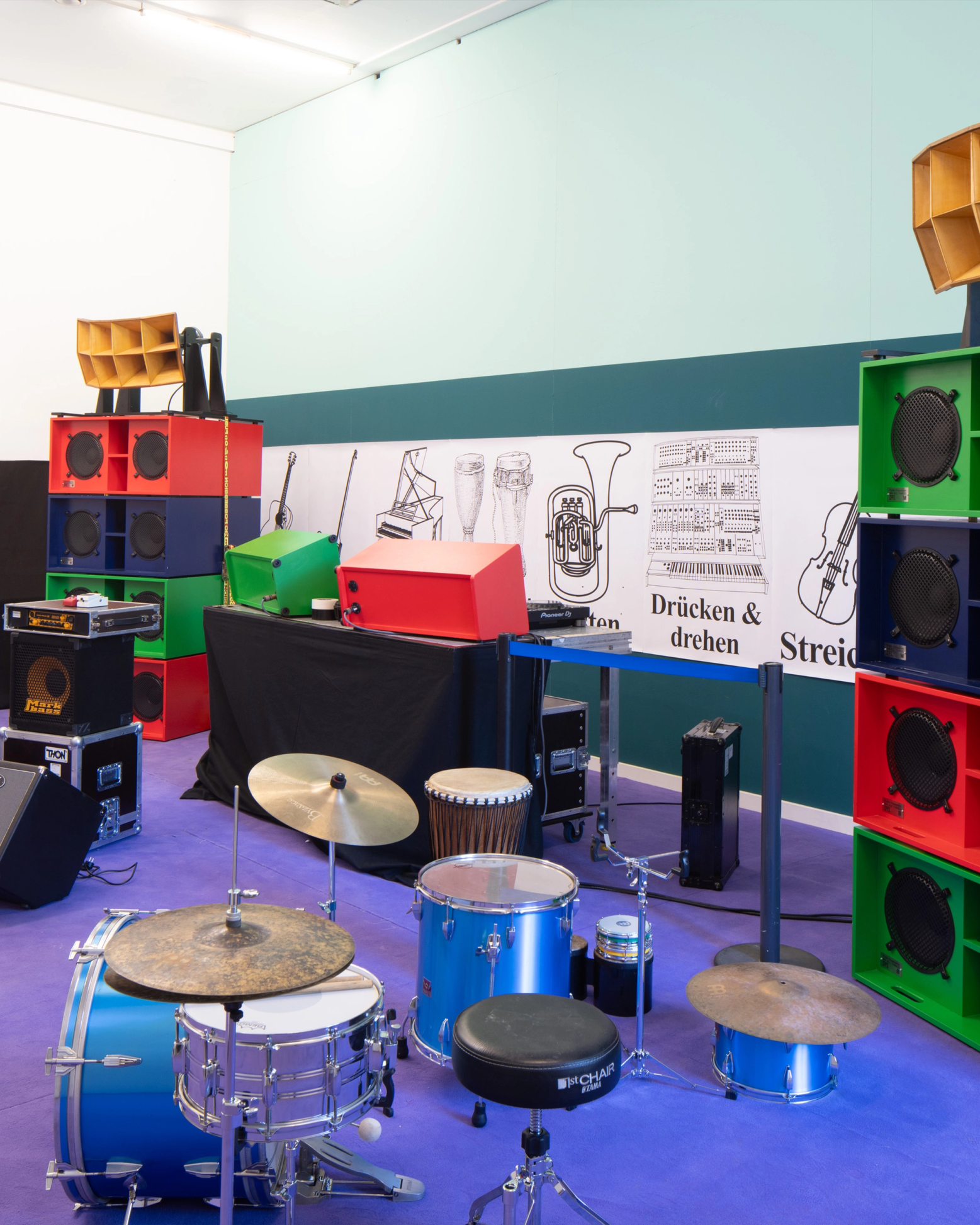 Stacks of colorful red, blue, and green speakers surround a blue drum set and cymbals on a purple floor, with a wall mural of musical instruments in the background.