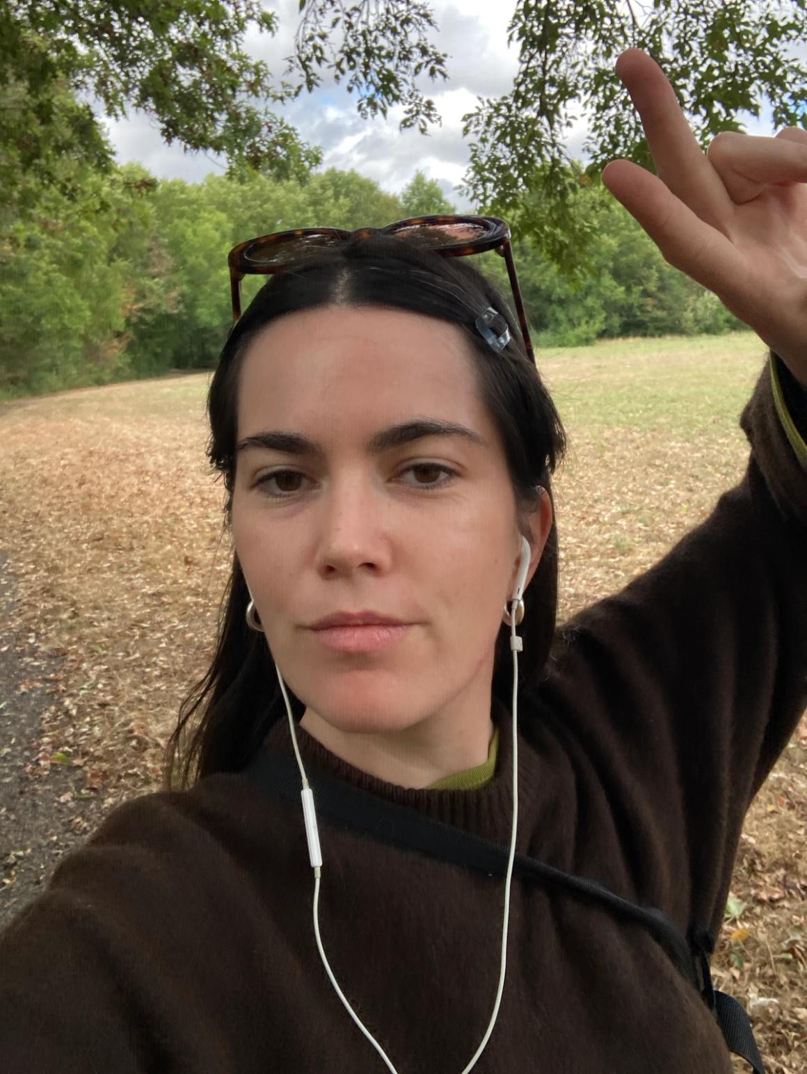 Woman wearing earbuds takes a selfie in a park, making a peace sign.