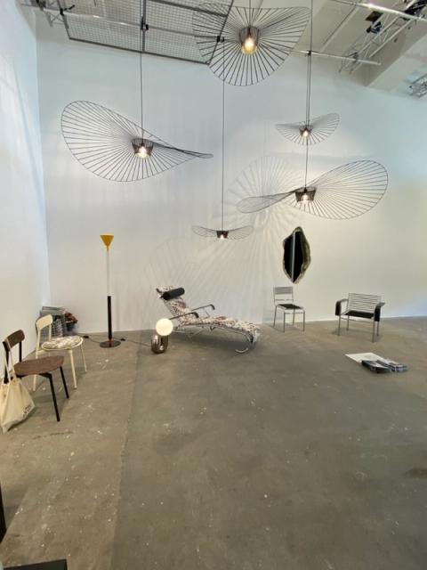 A showroom with several large, hat-like pendant lights, a lounge chair, assorted modern chairs, and a wall mirror.