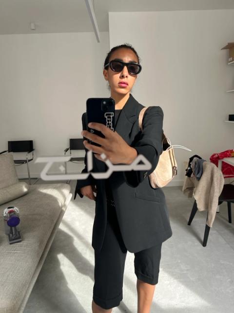Mirror selfie of a person in black sunglasses, blazer, and shorts, with a light shoulder bag and white Arabic text overlay.