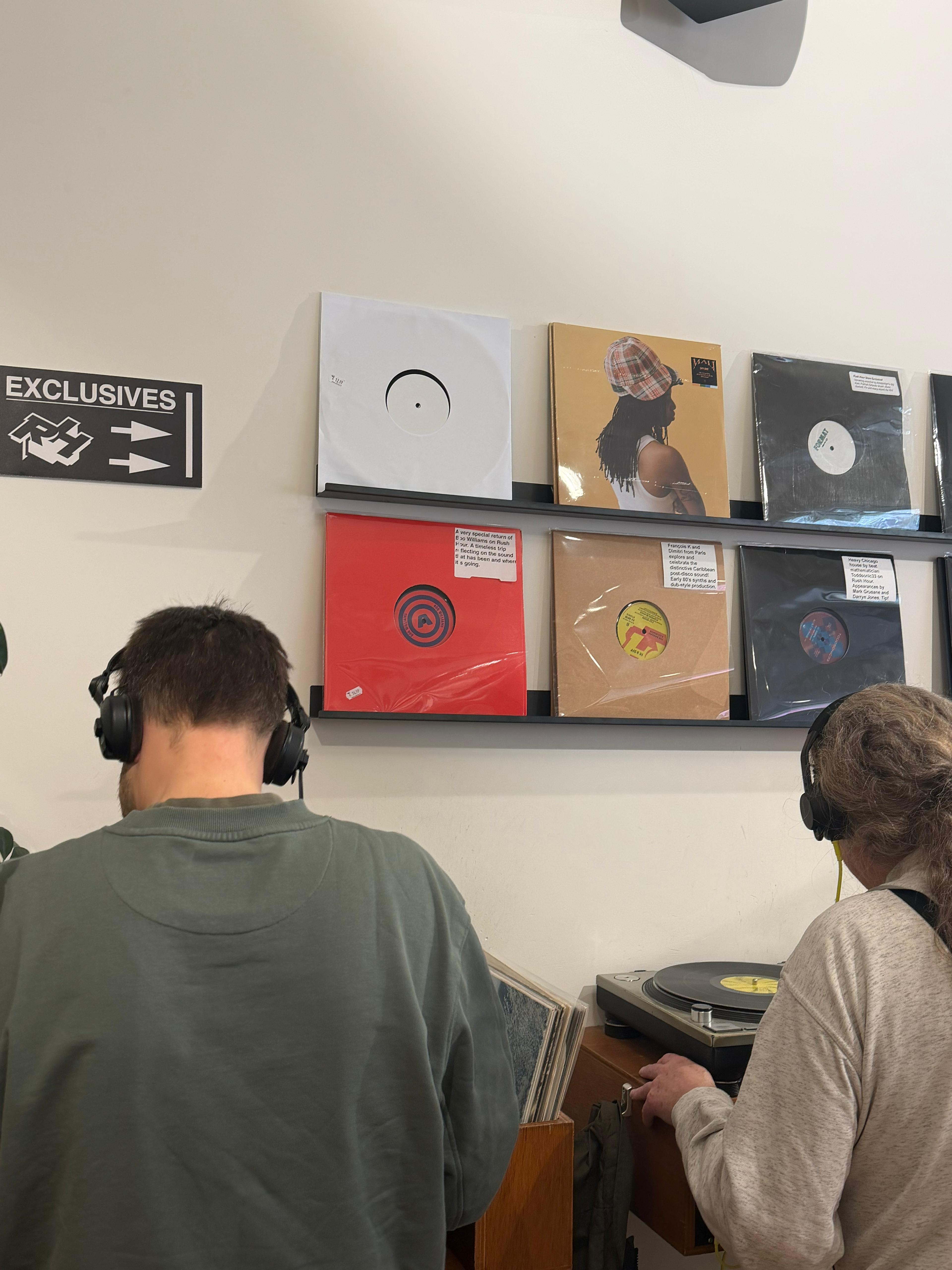 Two people wearing headphones at a record store, one using a turntable, with vinyl albums displayed on shelves.