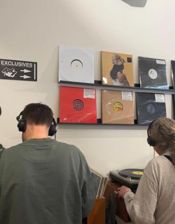 Two people wearing headphones at a record store, one using a turntable, with vinyl albums displayed on shelves.