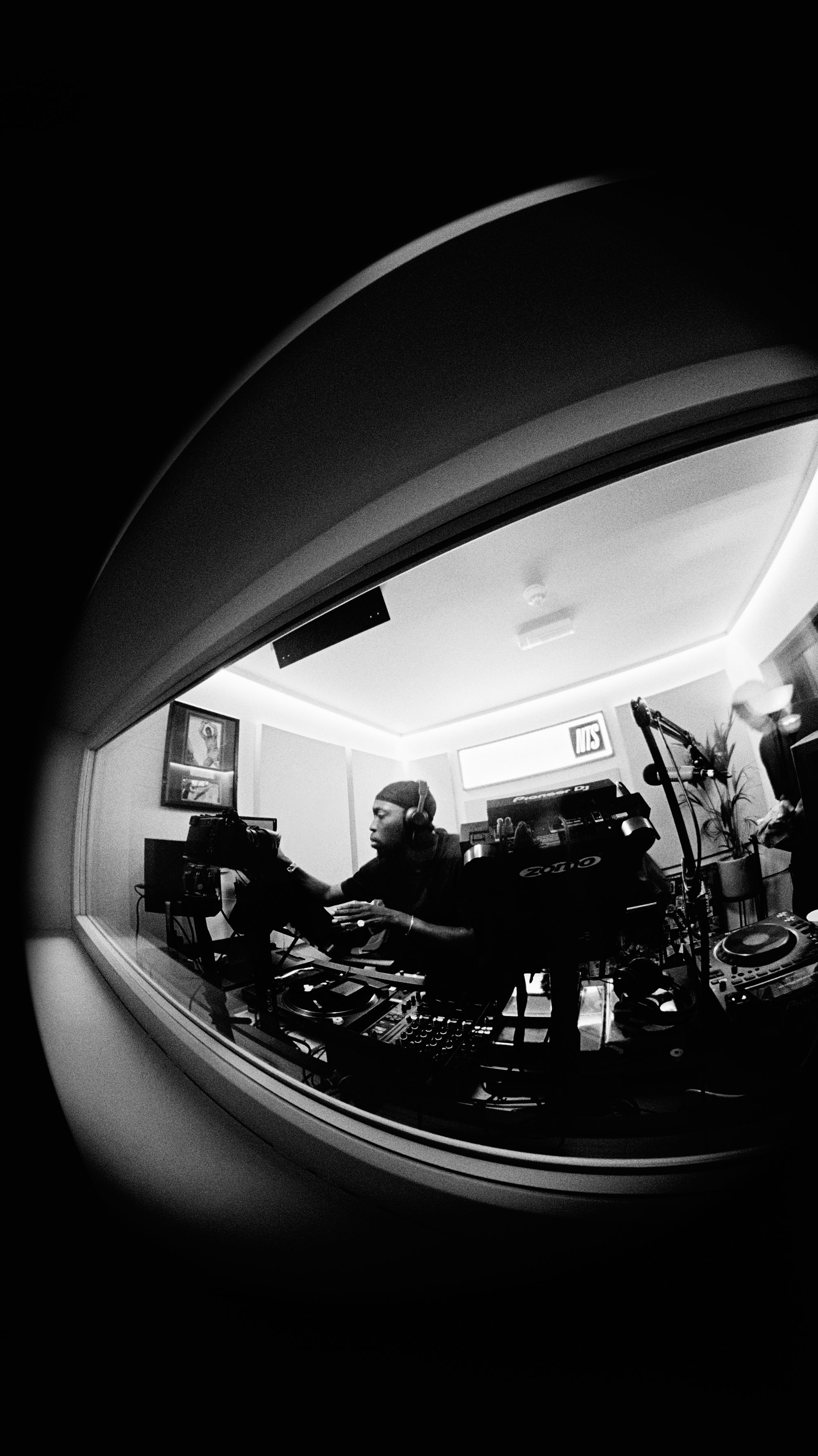 Black and white fisheye view of a DJ with headphones at a mixing console in a studio, seen through a window.