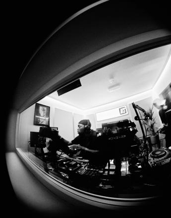 Black and white fisheye view of a DJ with headphones at a mixing console in a studio, seen through a window.