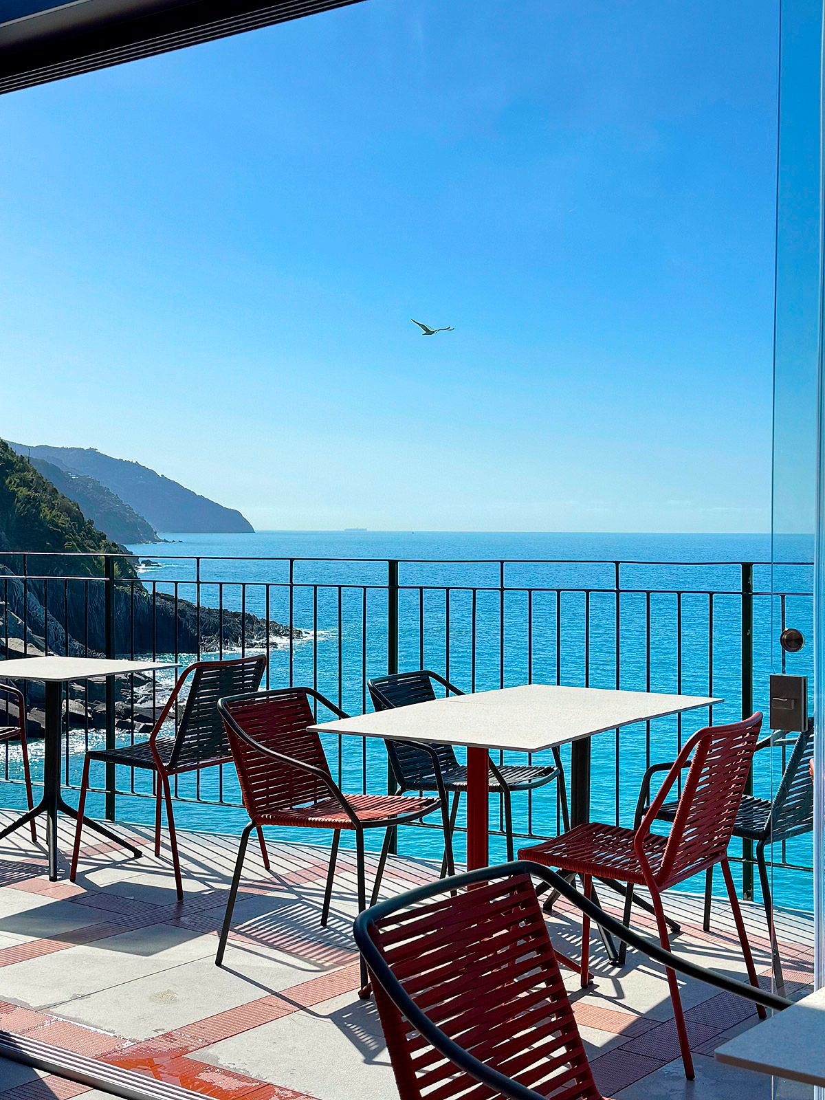 A cafe patio with red and black chairs overlooks a vibrant blue ocean and distant green coastline under a clear sky, with a bird flying overhead.