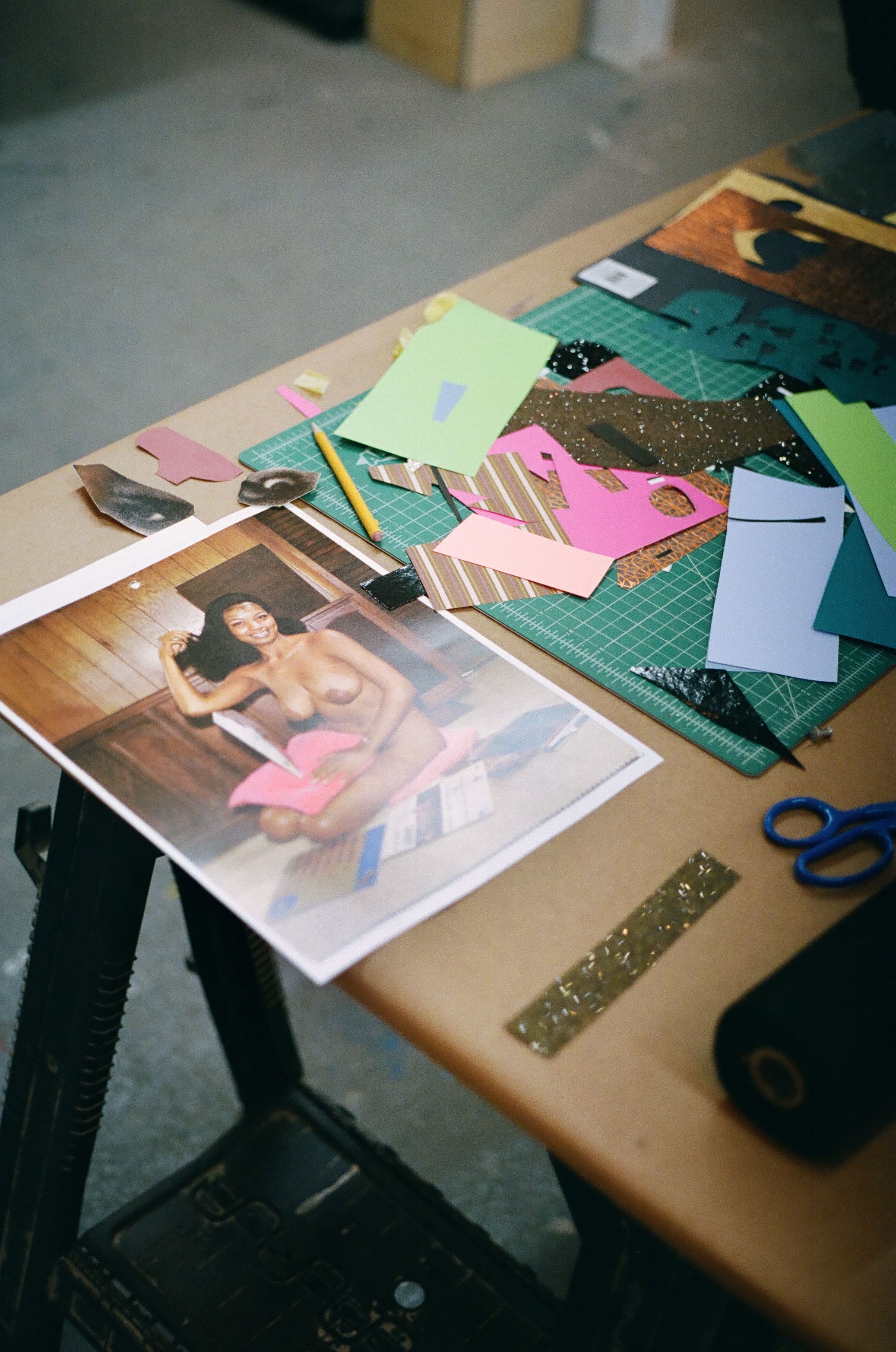 A craft table with a large print of a smiling nude Black woman, surrounded by colorful paper cutouts, a cutting mat, pencil, and scissors.