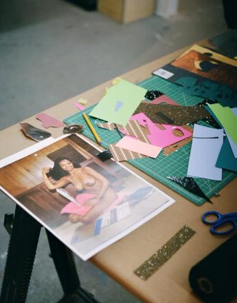 A craft table with a large print of a smiling nude Black woman, surrounded by colorful paper cutouts, a cutting mat, pencil, and scissors.