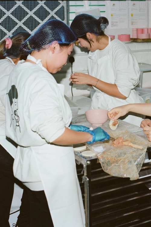 Workers in a food preparation area wearing hairnets and aprons, assembling dumplings with dough and meat filling.