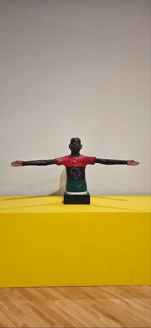 A sculpture of a dark-skinned figure with outstretched arms, wearing a red, black, and green shirt with an outline of Africa, on a yellow surface.