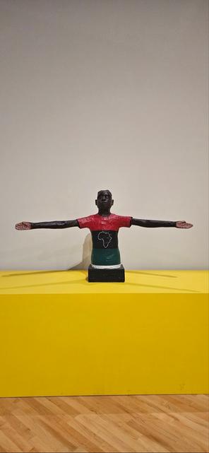 A sculpture of a dark-skinned figure with outstretched arms, wearing a red, black, and green shirt with an outline of Africa, on a yellow surface.