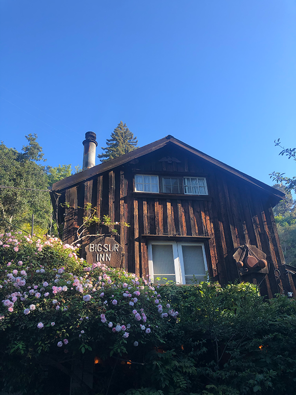 Rustic wooden Big Sur Inn building surrounded by pink roses and greenery under a clear blue sky.