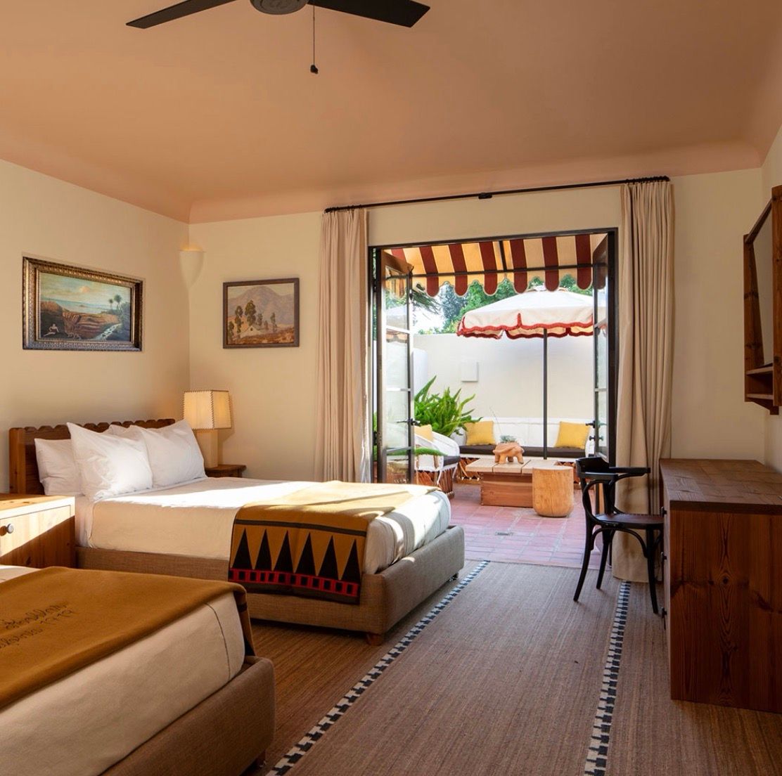 A warm-toned room with two beds and patterned blankets opens onto a sunny patio with an umbrella, awning, and outdoor furniture.