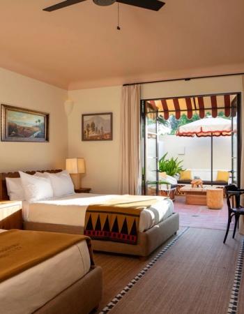 A warm-toned room with two beds and patterned blankets opens onto a sunny patio with an umbrella, awning, and outdoor furniture.