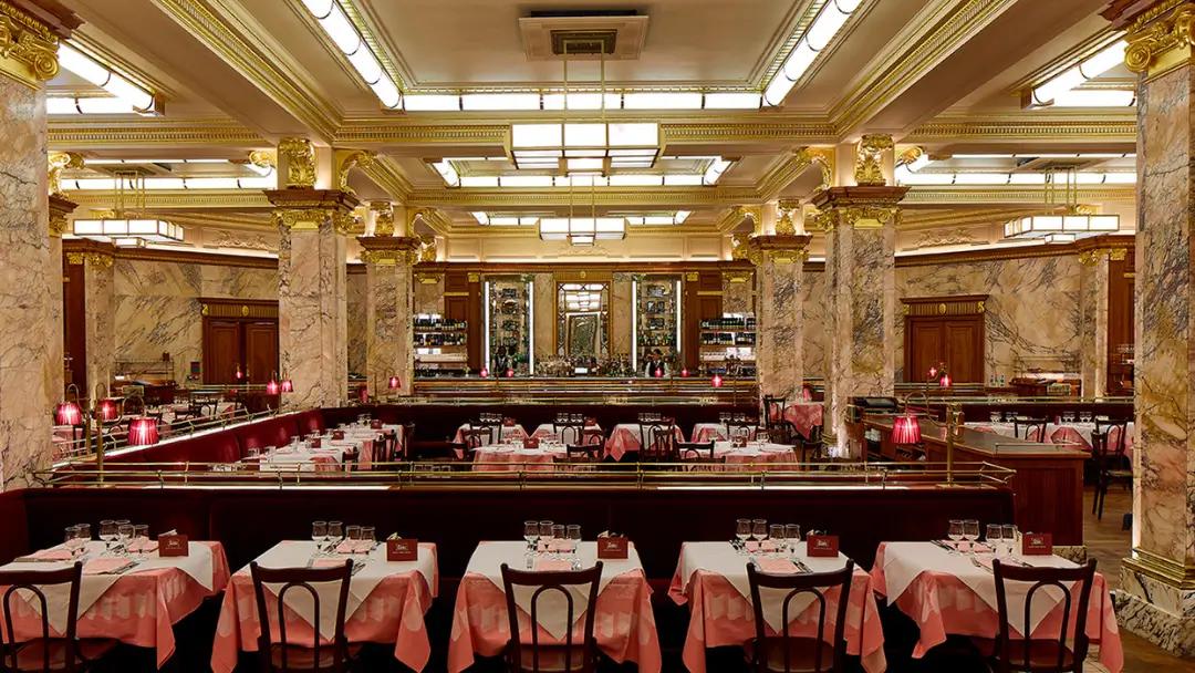An opulent restaurant interior with marble columns, gold trim, red velvet banquettes, and empty tables set with pink and white checkered tablecloths.