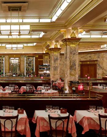 An opulent restaurant interior with marble columns, gold trim, red velvet banquettes, and empty tables set with pink and white checkered tablecloths.