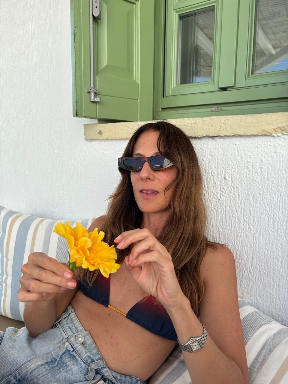 A woman in sunglasses and a bikini top holds a yellow flower, leaning on striped cushions with green shutters behind her.