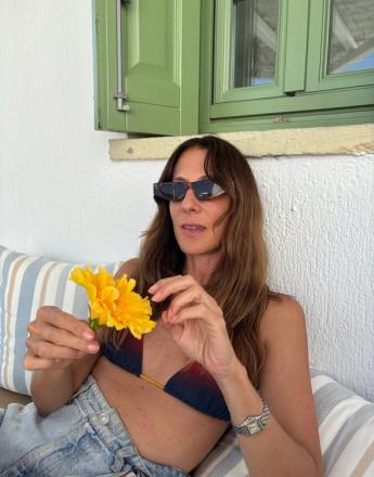 A woman in sunglasses and a bikini top holds a yellow flower, leaning on striped cushions with green shutters behind her.