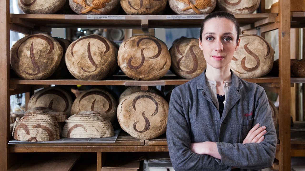 A woman with crossed arms stands in front of shelves filled with round loaves of bread scored with stylized P and S designs.