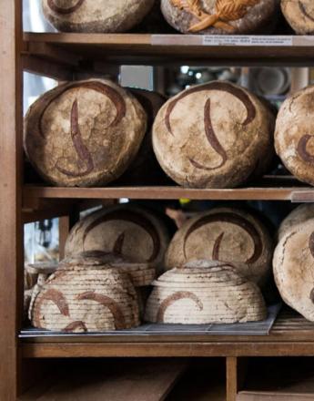 A woman with crossed arms stands in front of shelves filled with round loaves of bread scored with stylized P and S designs.