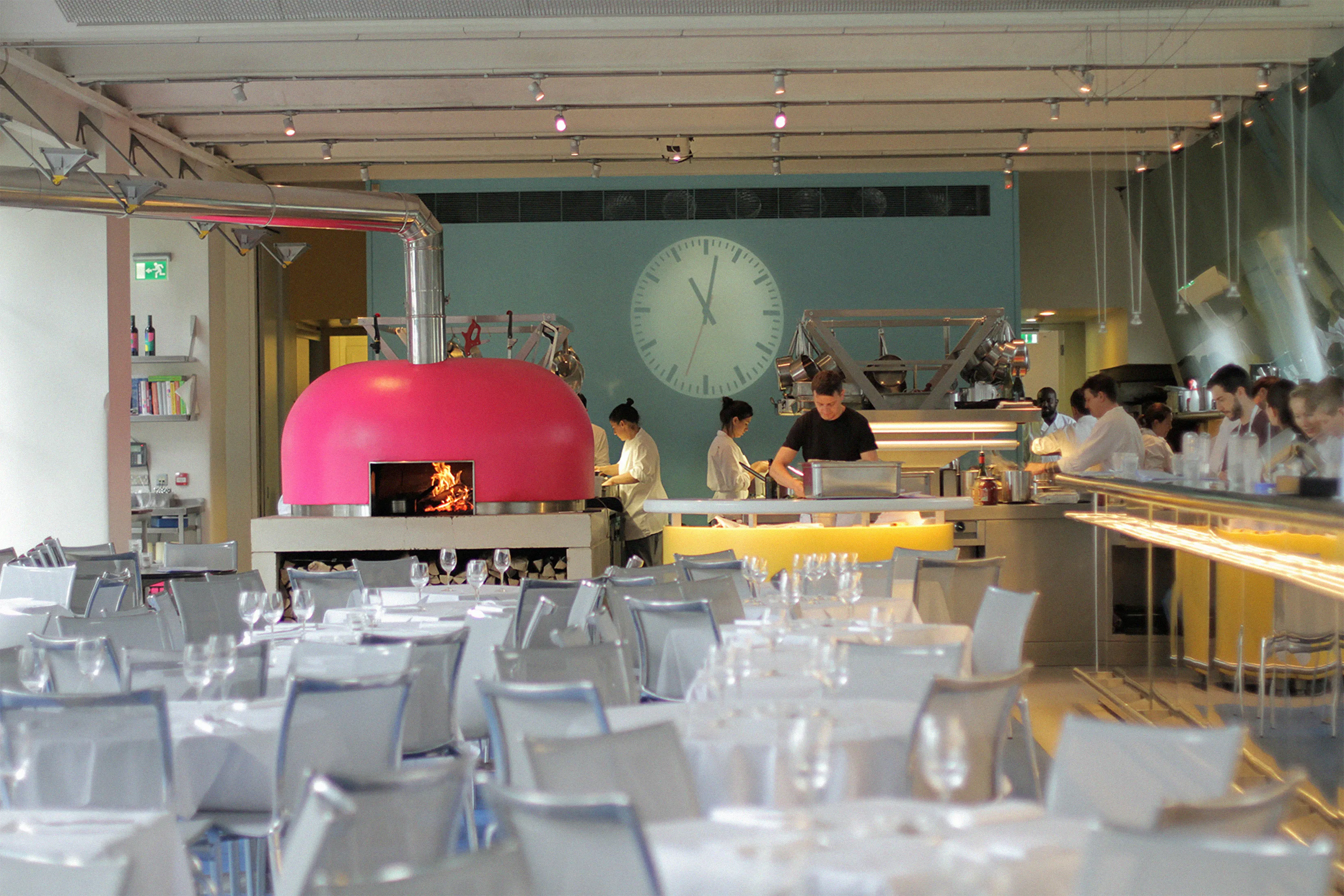 Modern restaurant dining room with white-clothed tables, grey chairs, and a bar with red flowers.