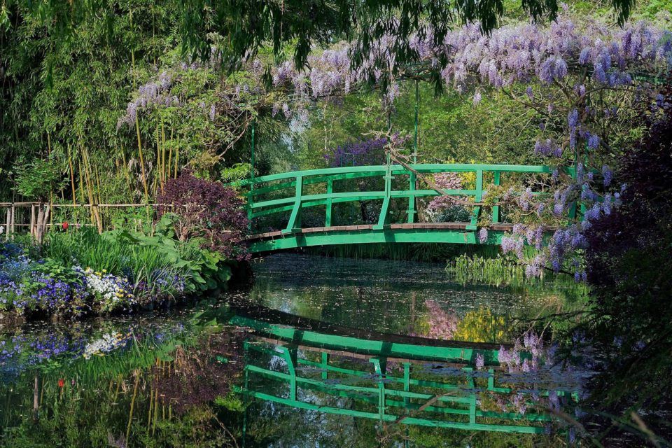 A green Japanese bridge over a pond surrounded by purple wisteria and lush foliage, with reflections in the water.