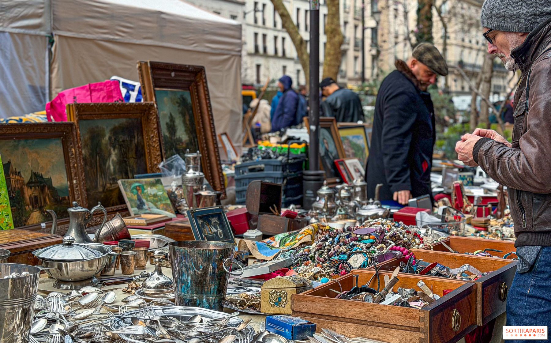 A bustling outdoor flea market with a man browsing antique silverware, paintings, and jewelry.