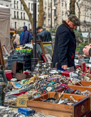 A bustling outdoor flea market with a man browsing antique silverware, paintings, and jewelry.