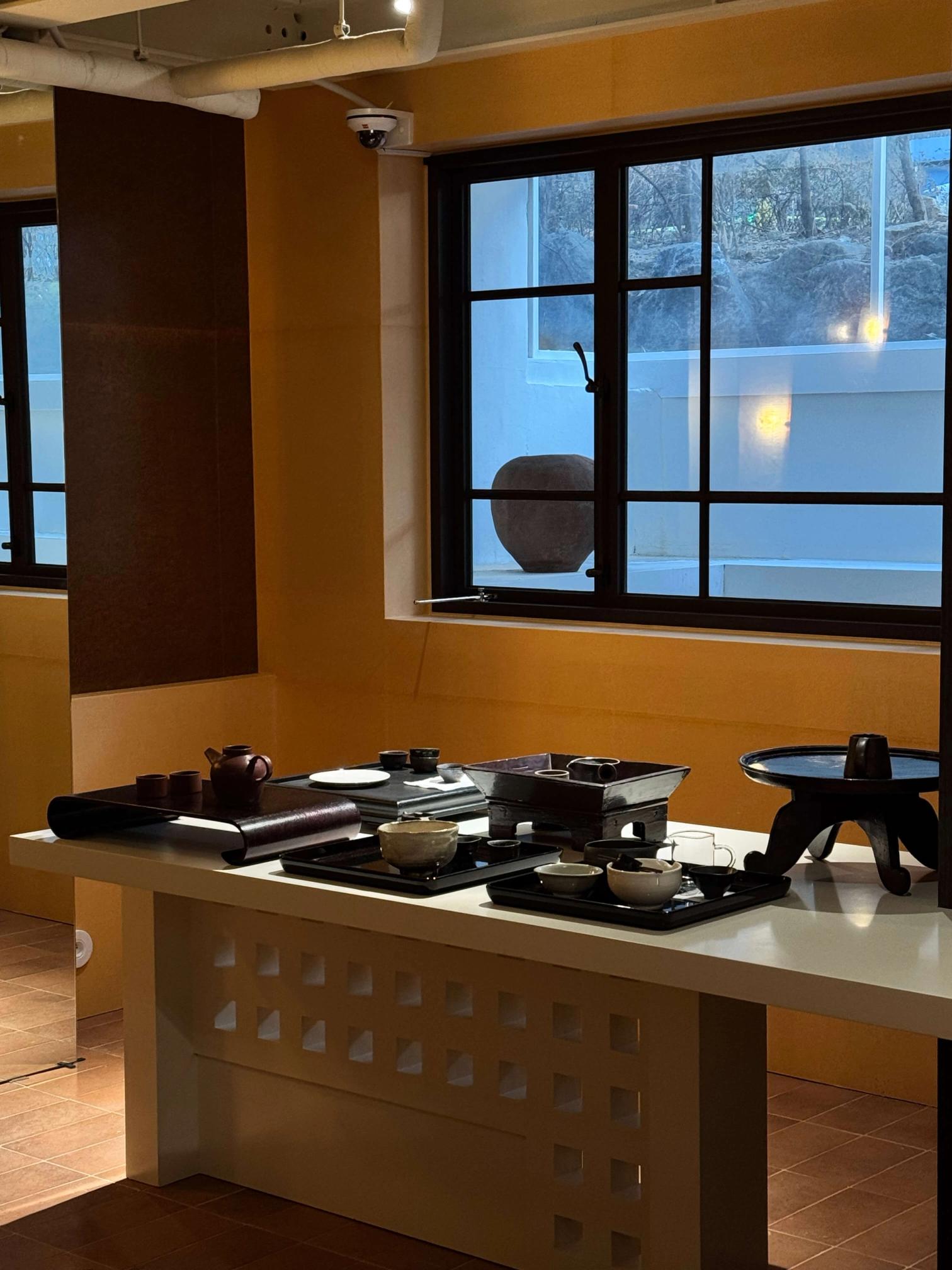 A white table displays multiple dark trays and traditional tea ceremony sets in a bright room with orange walls and large windows.