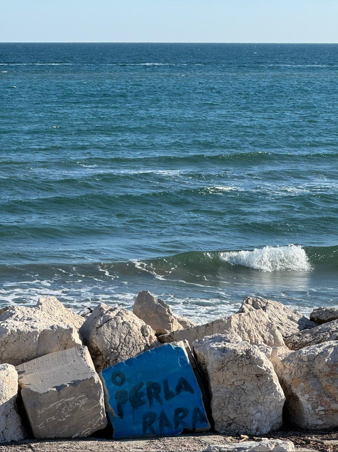 Ocean waves lap against a rocky shore, where a blue concrete block is painted with 'O PERLA RARA'.