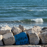 Ocean waves lap against a rocky shore, where a blue concrete block is painted with 'O PERLA RARA'.