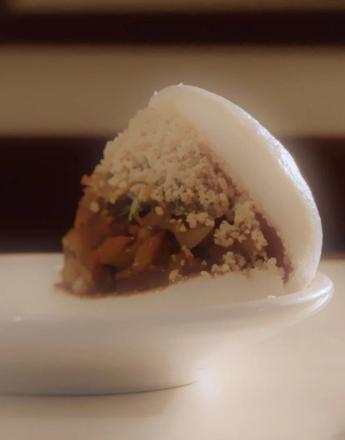An open bao bun filled with savory ingredients and topped with white crumbs, served on a white dish.