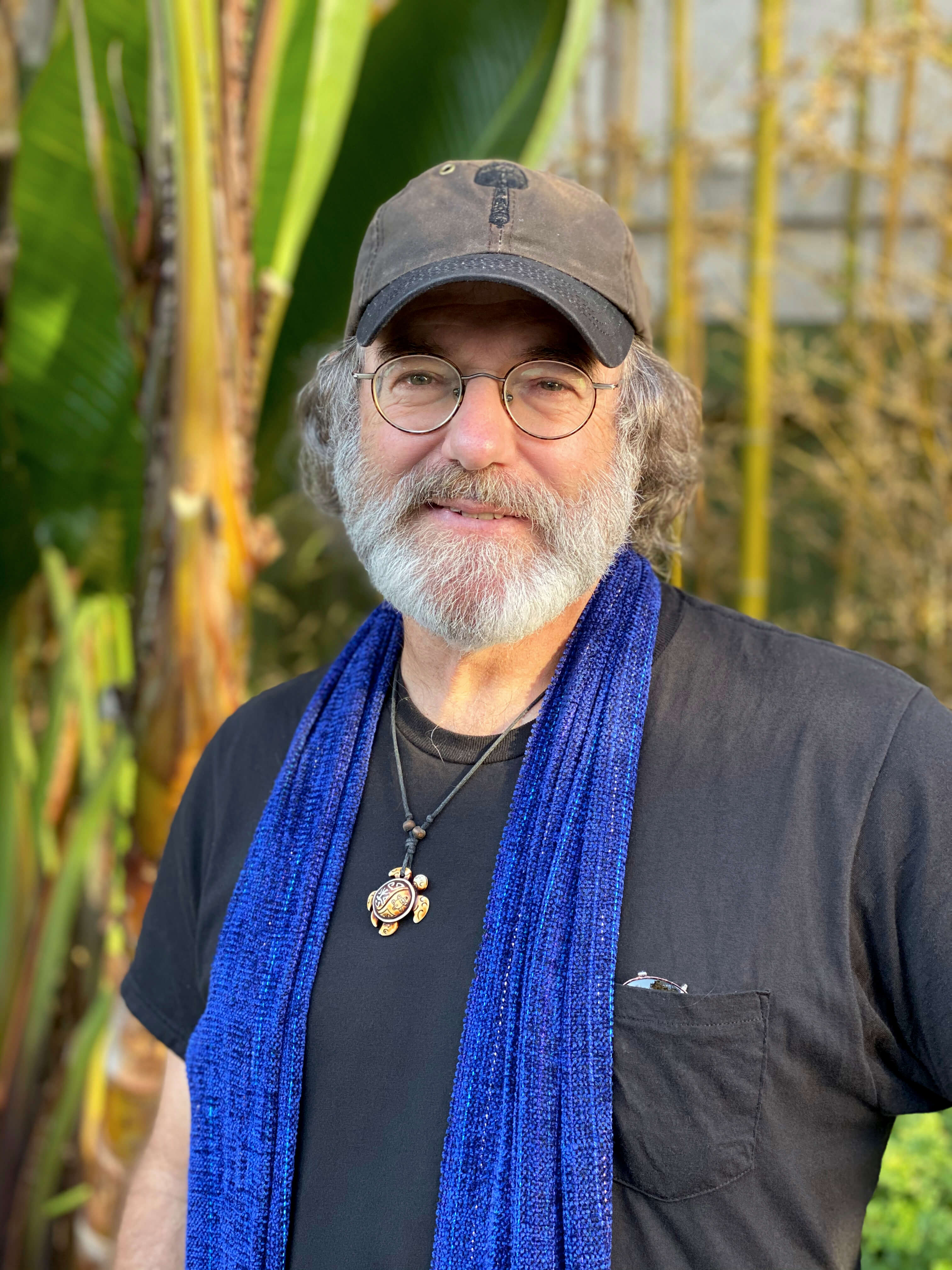 A smiling man with a grey beard, round glasses, and a cap with a mushroom logo, wearing a blue scarf and a turtle necklace, stands among tropical plants.