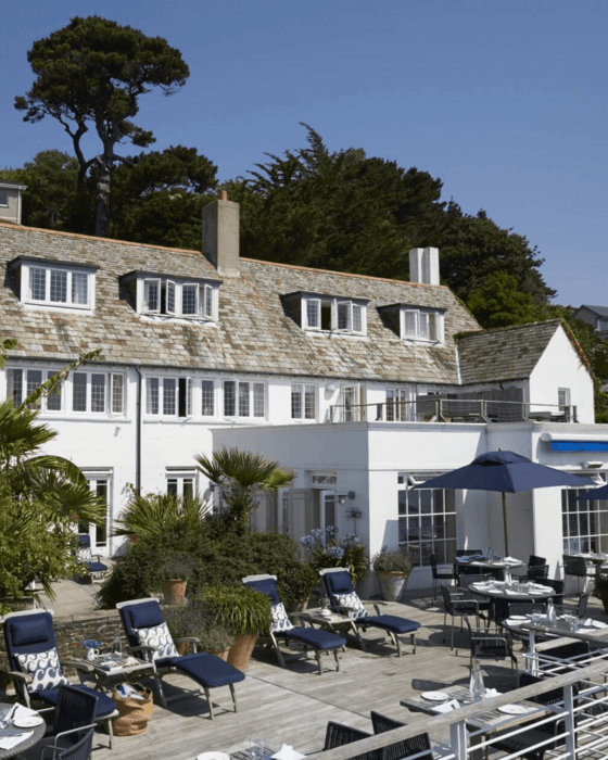 A white building with a slate roof and dormer windows, fronting a sunny wooden deck with blue sun loungers, dining tables, and blue umbrellas.