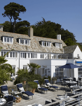 A white building with a slate roof and dormer windows, fronting a sunny wooden deck with blue sun loungers, dining tables, and blue umbrellas.