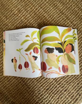 Open children's book showing colorful illustrations of people and large seeds growing into plants, on a woven mat.