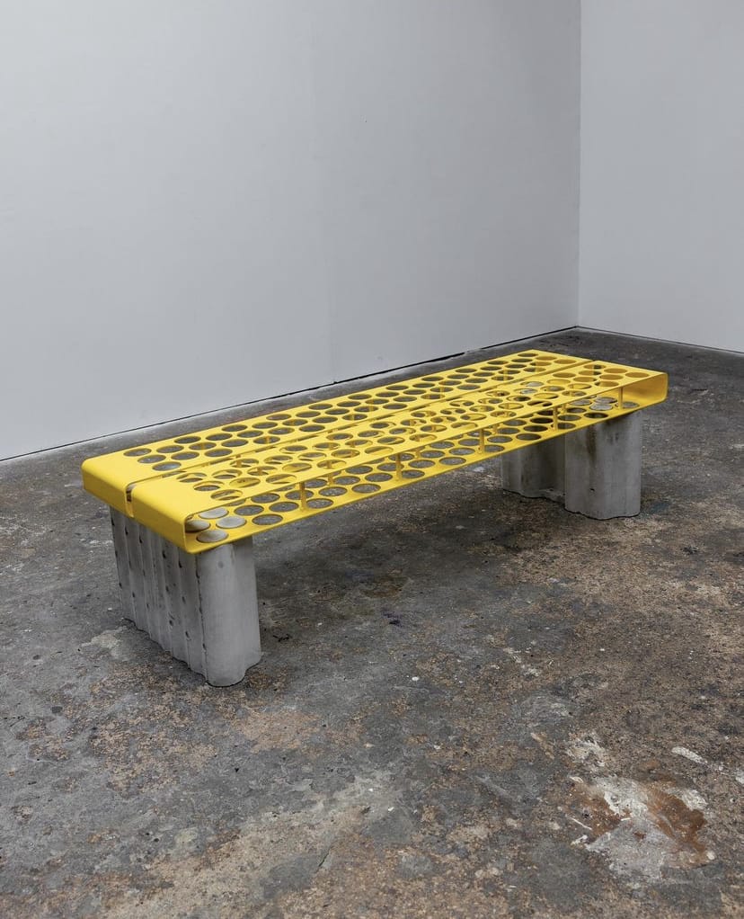 A bright yellow perforated metal bench with concrete legs, coins visible in its holes, in a sparse room.