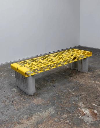 A bright yellow perforated metal bench with concrete legs, coins visible in its holes, in a sparse room.