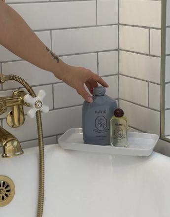 A hand reaches for a grey bottle, one of two on a white tray on the rim of a bathtub with gold fixtures and white tiled walls.