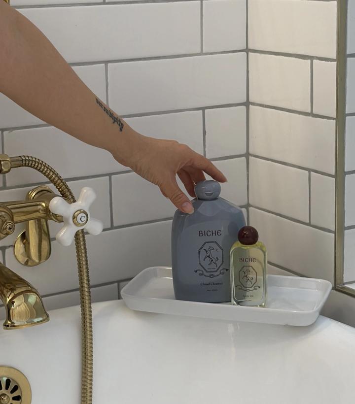 A hand reaches for a gray Biche cleanser bottle on a white tray in a bathtub.
