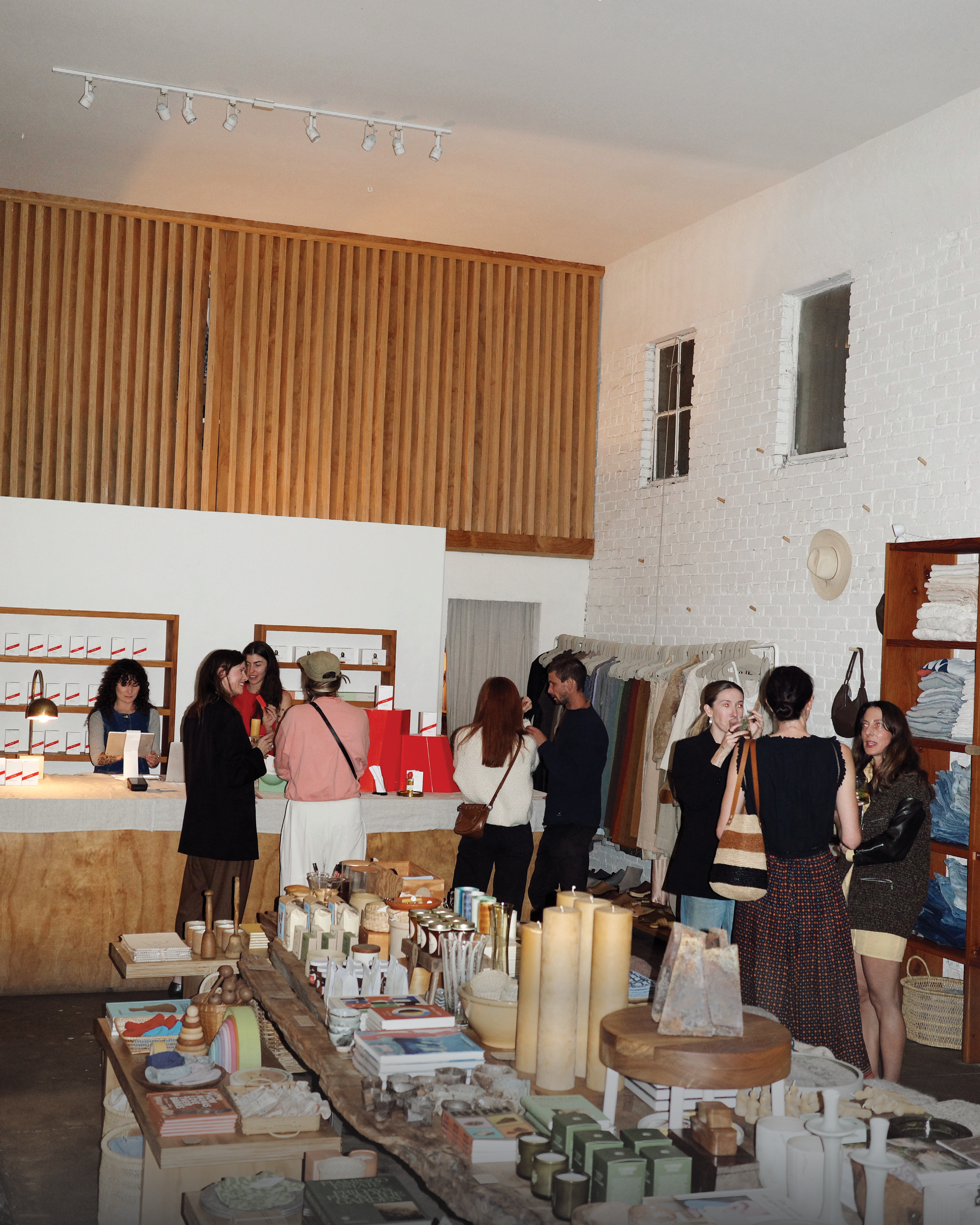 Inside a bustling modern boutique with white walls and wood accents, people browse clothing racks and rustic tables filled with candles, books, and home goods.